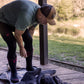 A man outdoors on a wooden deck adjusts black knee-high Merino socks while wearing a green LOC T-shirt, LOC black cap, and black Merino leggings. A pair of dark socks or a garment lies on the deck beside him, with grass and trees in the background.