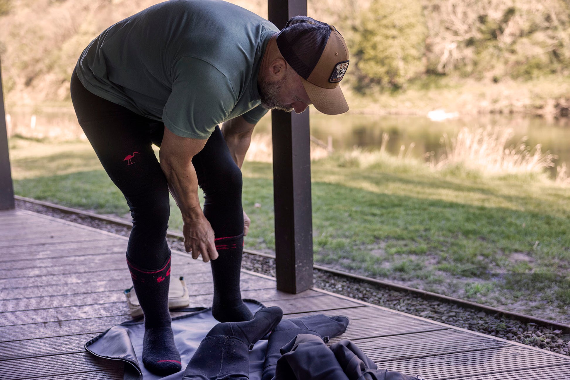 A man outdoors on a wooden deck adjusts black knee-high Merino socks while wearing a green LOC T-shirt, LOC black cap, and black Merino leggings. A pair of dark socks or a garment lies on the deck beside him, with grass and trees in the background.
