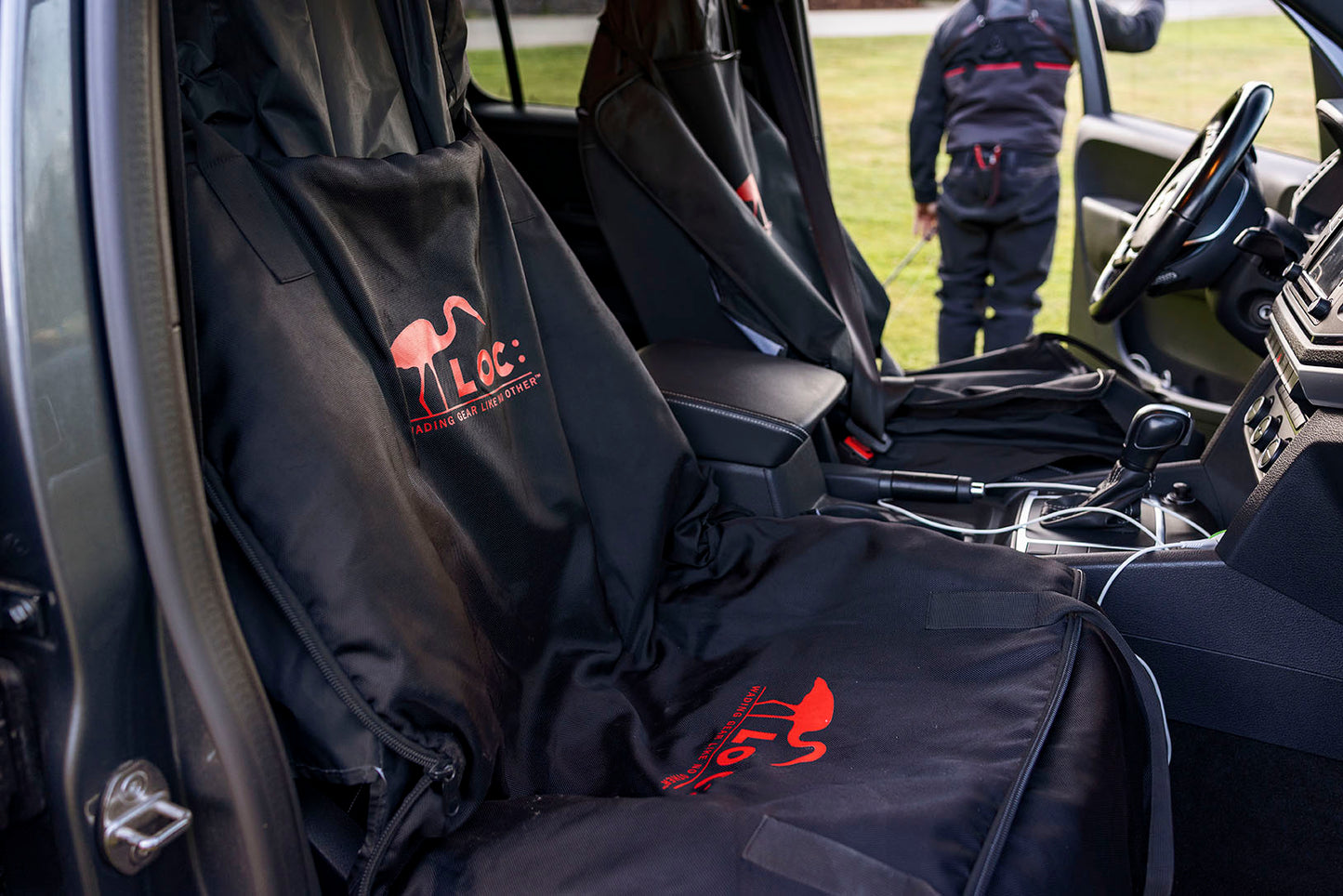 Black LOC 3-in-1 car seat covers with red LOC logos and text, shown installed on the front seats of a vehicle interior. A person is visible standing outside the open driver's side door on grass in the background. He´s dressed in LOC Waders.