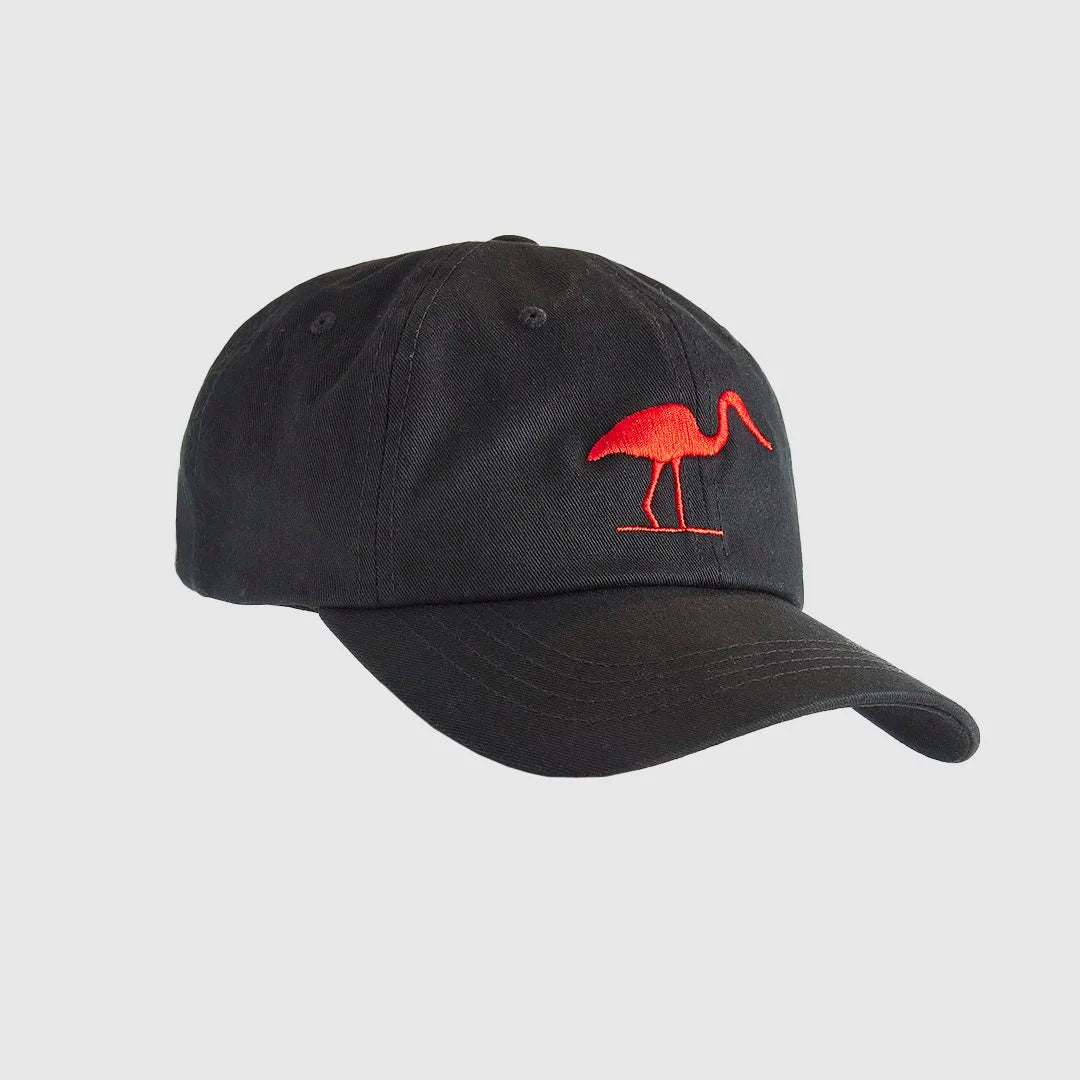 A black low profile dad cap with a red stitched heron symbol on the front sitting on top of a tree stump.