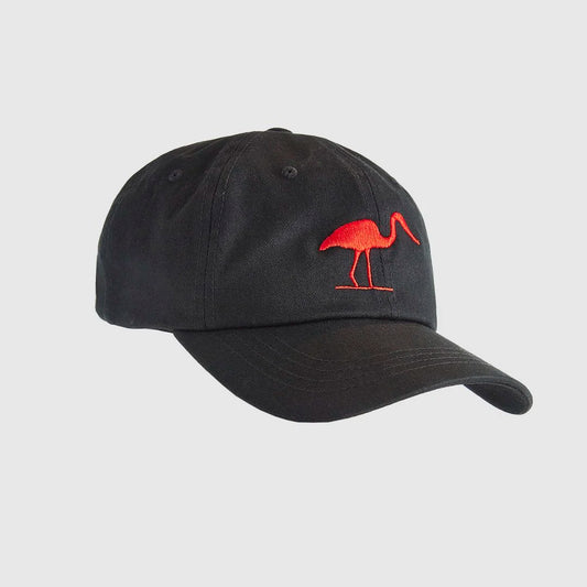 A black low profile dad cap with a red stitched heron symbol on the front sitting on top of a tree stump.